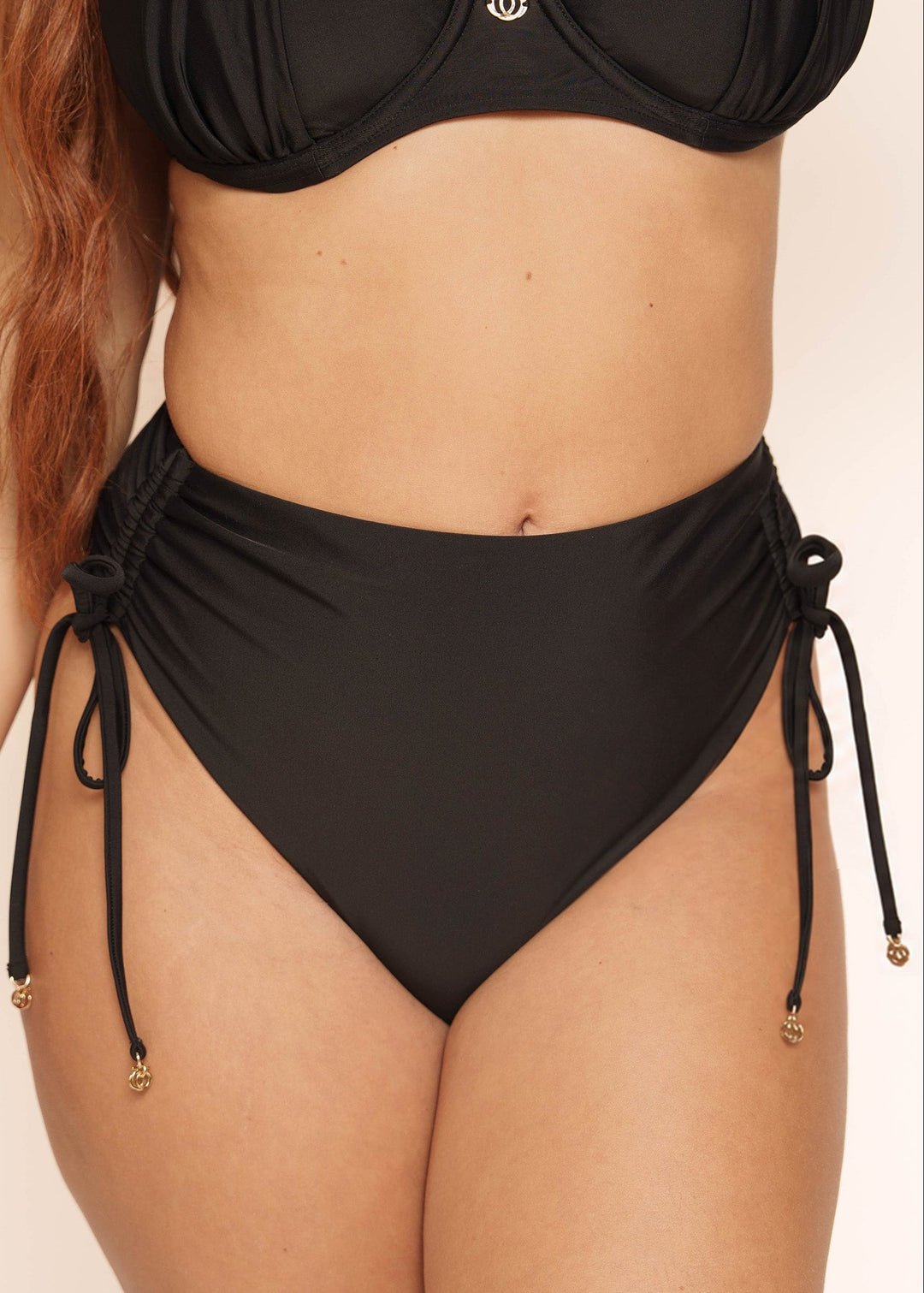 A model wearing a black two-piece bikini with high-waisted side-tie bottoms and a matching bandeau-style top, shown against a plain light beige studio background.