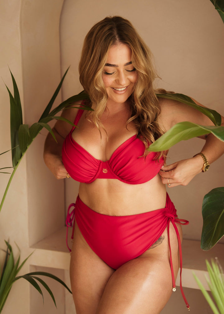 A woman modeling a bright red two-piece bikini with side-tie bottoms, standing in a warm beige architectural setting with tropical green leaves framing the scene.