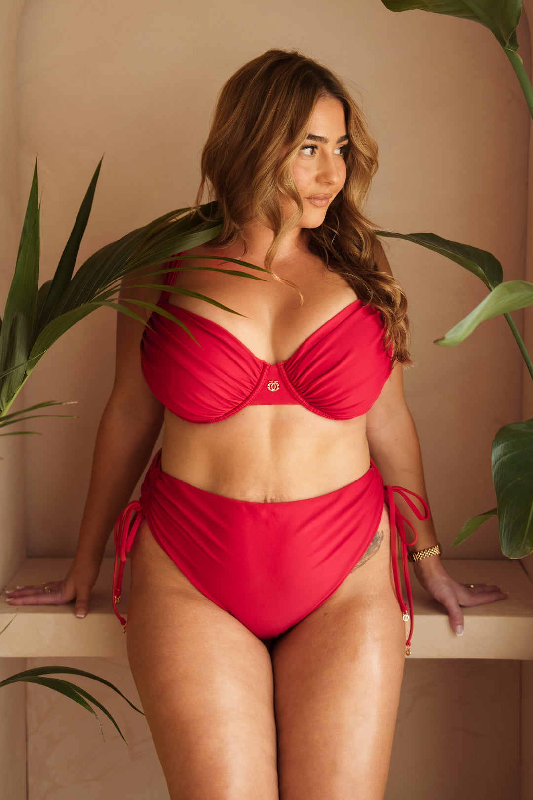 A woman modeling a bright red two-piece bikini with a bandeau-style top and high-waisted side-tie bottoms, posed against a warm beige wall with tropical green leaves framing the scene.